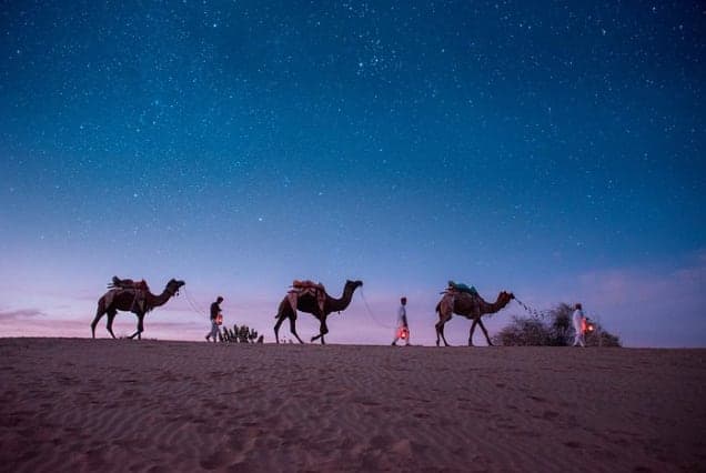 Camels and their handlers crossing sand dunes under a starry sky at dusk