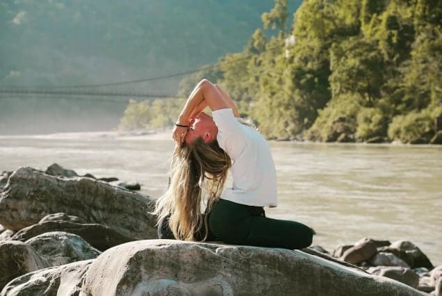 A woman in a seated yoga backbend on riverside rocks, with a suspension bridge and forested hills across the water