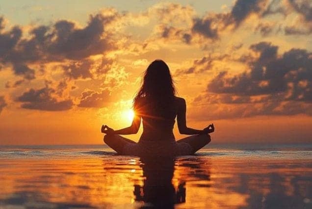 Silhouette of a woman meditating cross-legged at the water's edge against a golden sunset
