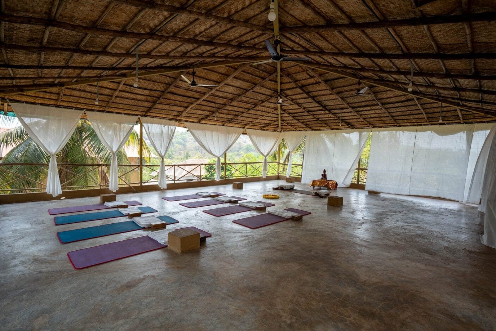 An open-sided yoga hall with a thatched roof, mats laid out and white curtains facing the trees