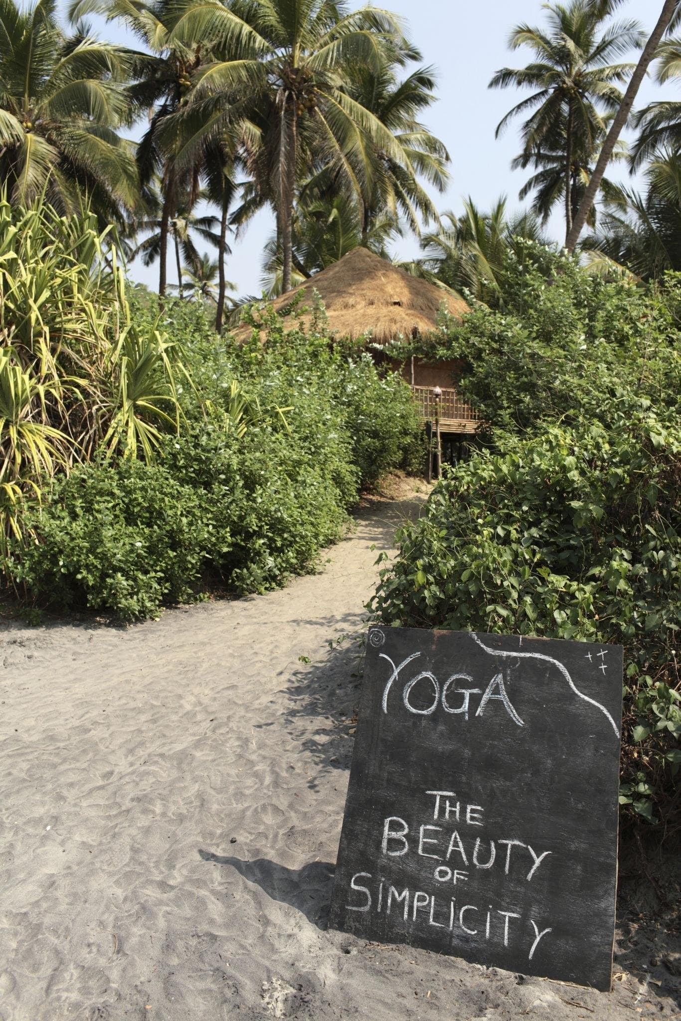 A sandy path through palms to a thatched hut, with a chalkboard sign reading 'Yoga: the beauty of simplicity'