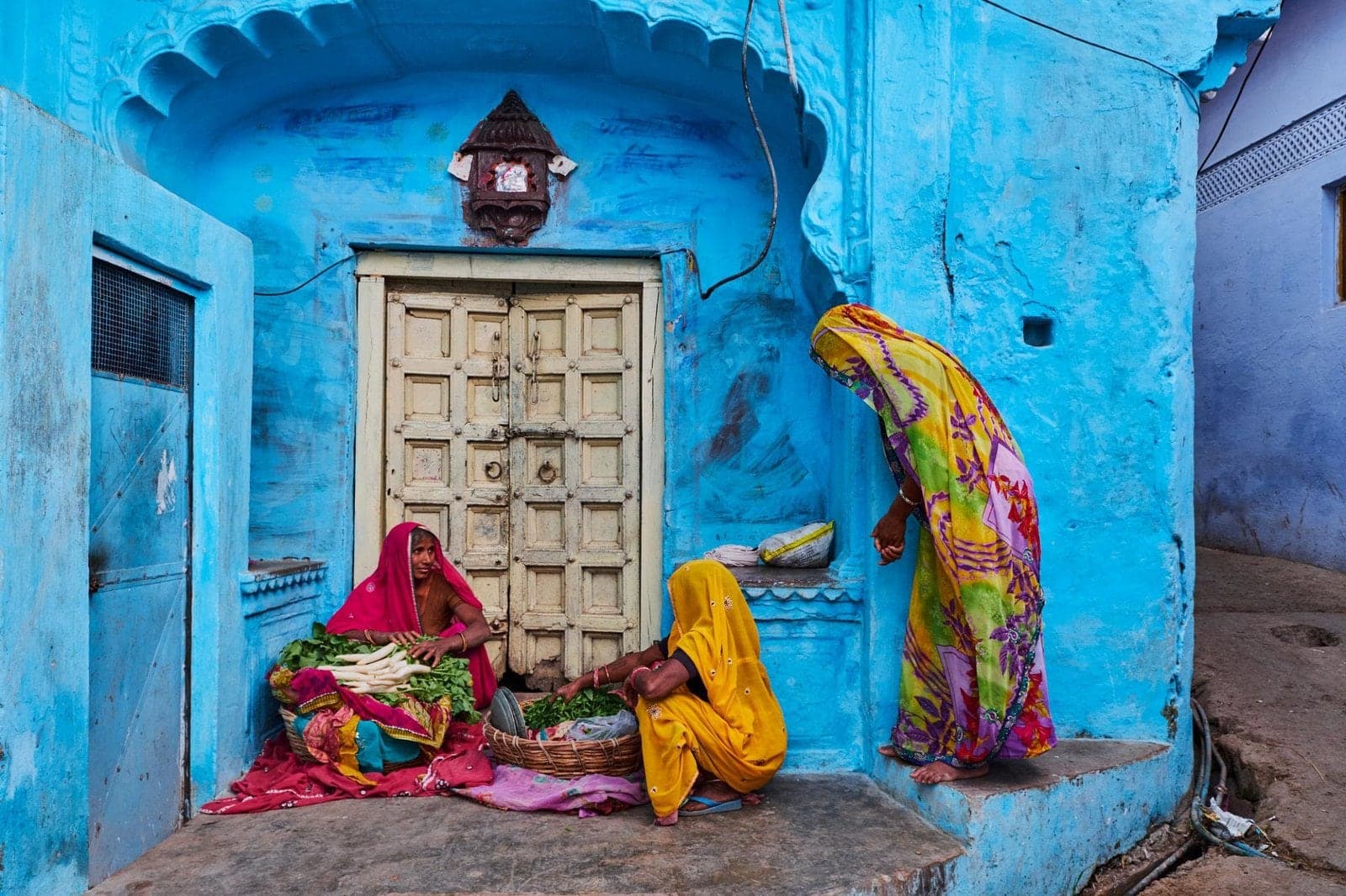 Women in bright saris selling vegetables beside a carved door in a blue-painted lane of Jodhpur