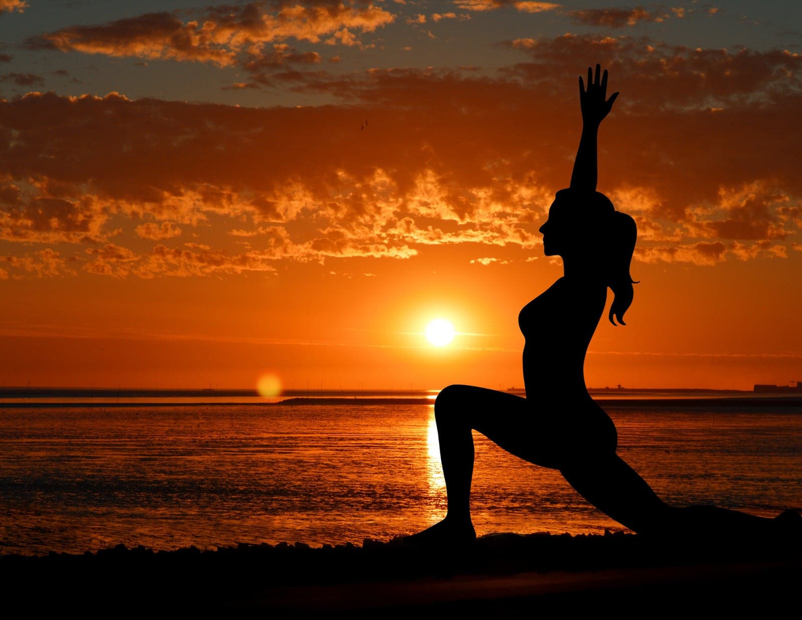 Silhouette of a woman in a high lunge yoga pose against an orange sunset over the sea