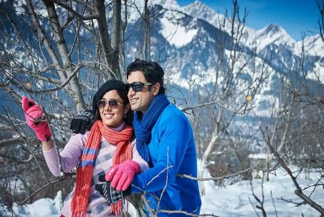A couple in winter scarves and gloves taking a photo among snow-covered trees and Himalayan peaks