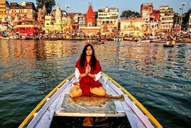 A woman in red meditating in a wooden boat on the Ganges, facing the ghats of Varanasi