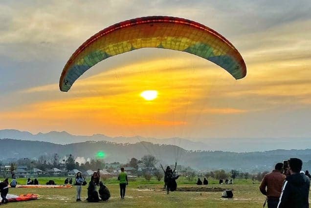 A paraglider launching into the sunset above a meadow where pilots and onlookers gather
