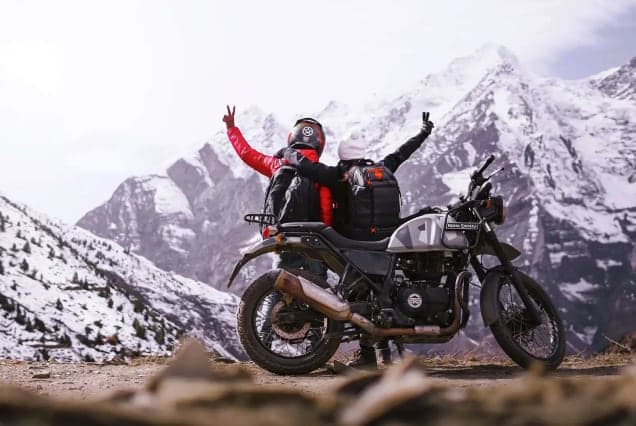 A rider and passenger on a Royal Enfield Himalayan raising their arms in front of snow-capped peaks