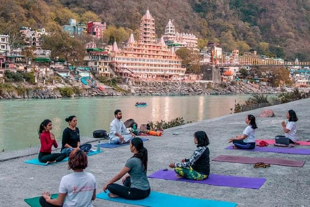 A group yoga class on mats beside the Ganges in Rishikesh, with the tiered Tera Manzil temple behind