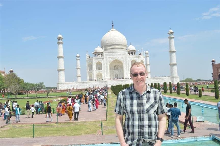 A traveller in a checked shirt smiling in front of the Taj Mahal, with visitors along the garden paths