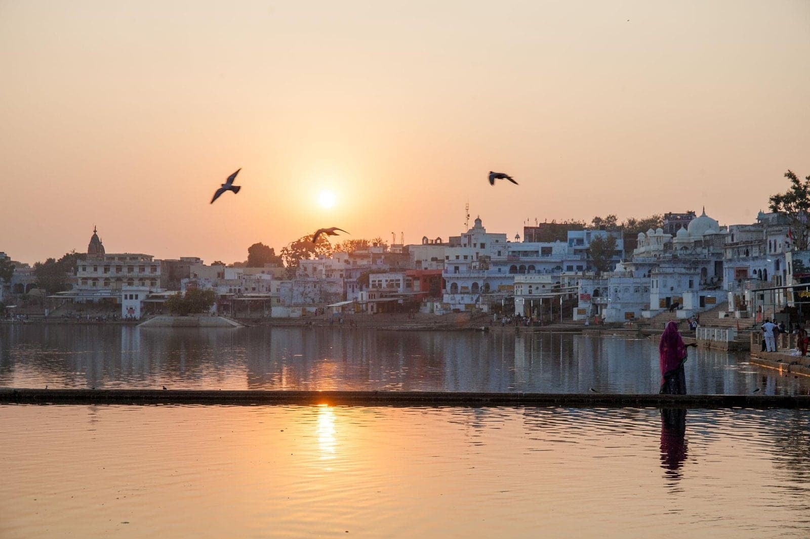Sunset over Pushkar Lake, with birds overhead and a woman walking along the ghats